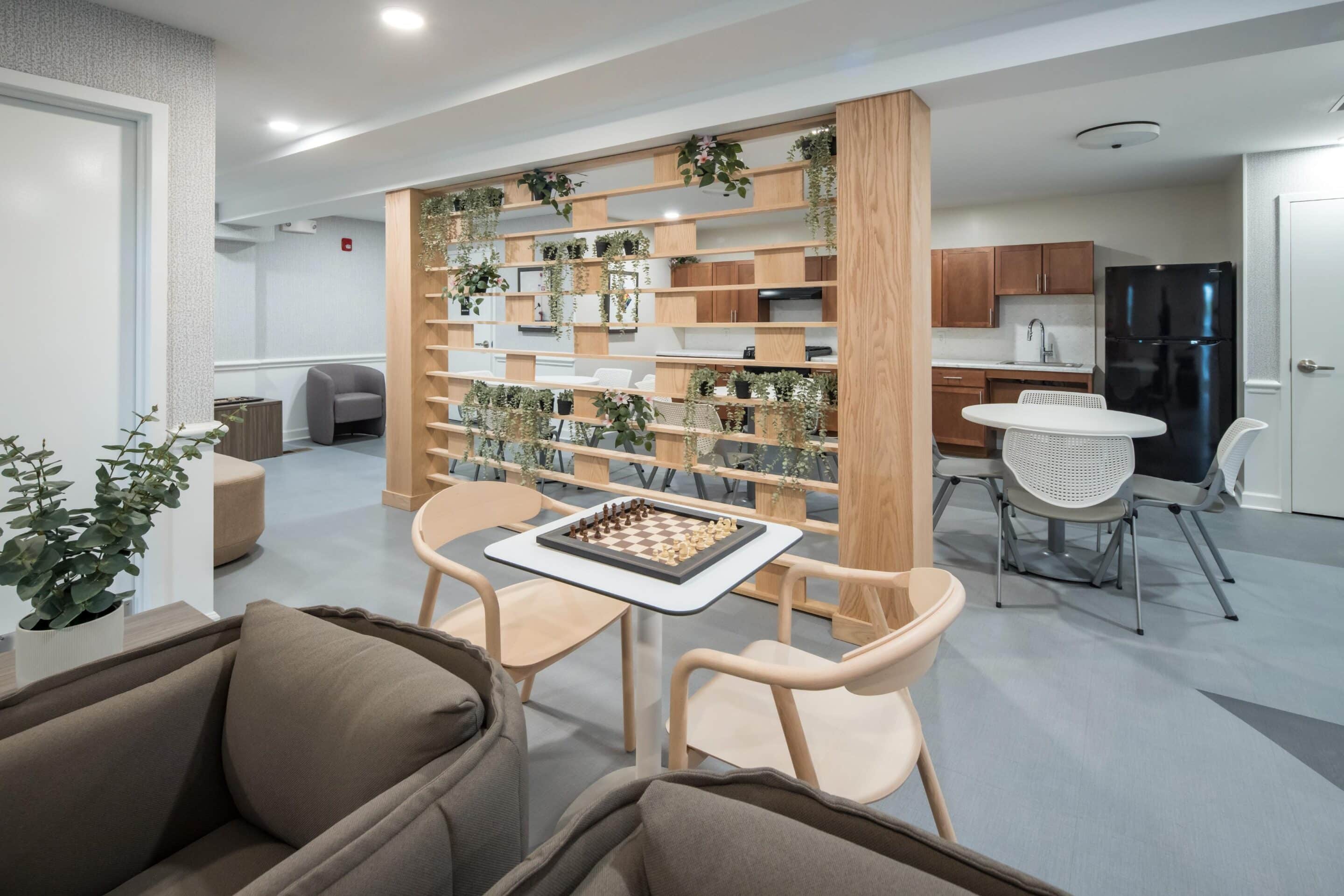 Modern lounge area at Ridgecrest Village, a Tax Credit Community, features a chessboard on a table, brown couches, open shelving with plants, dining tables, and a kitchenette with wooden cabinets and a black refrigerator.