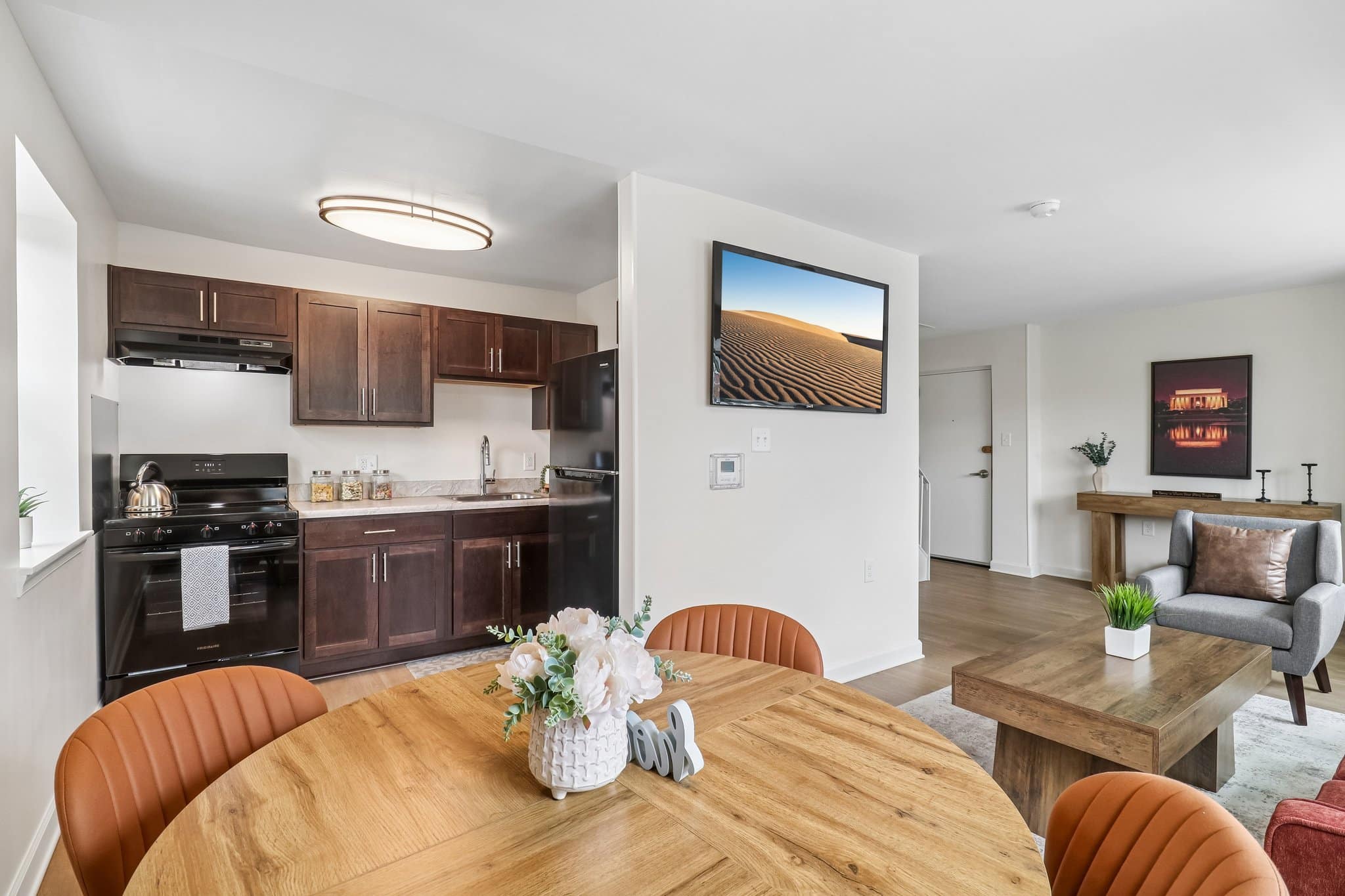 Open-plan apartment with a kitchen featuring dark cabinets, a dining table with orange chairs, and a living area with modern decor and neutral colors.
